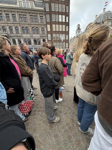 A crowd of people stands attentively on the cobblestone Dam Square in Centrum, Amsterdam, looking towards an unseen focal point